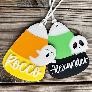 May include: Two Halloween-themed candy corn ornaments with personalized names. One is orange, yellow, and white with a ghost and the name "Rocco". The other is green, black, and white with a skull and the name "Alexander".