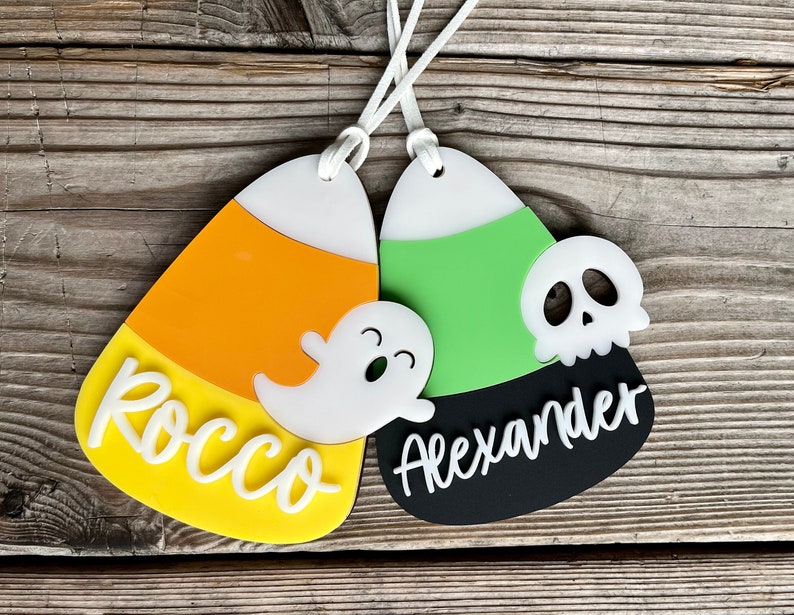 May include: Two Halloween-themed candy corn ornaments with personalized names. One is orange, yellow, and white with a ghost and the name "Rocco". The other is green, black, and white with a skull and the name "Alexander".