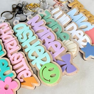 May include: A collection of personalized wooden keychains featuring layered acrylic names in various colors including teal, pink, lavender, and blue. Each keychain is accented with shapes like flowers, lightning bolts, smiley faces, bows, shells, and stars.