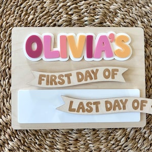 K&ouml;nnte beinhalten: Ein rechteckiges Holzschild mit dem Namen OLIVIA'S in erhabenen, bunten Buchstaben. Unter dem Namen sind zwei h&ouml;lzerne Banner-Ausschnitte angebracht, eines mit der Aufschrift FIRST DAY OF und das andere mit LAST DAY OF, &uuml;ber einer wei&szlig;en Platte.