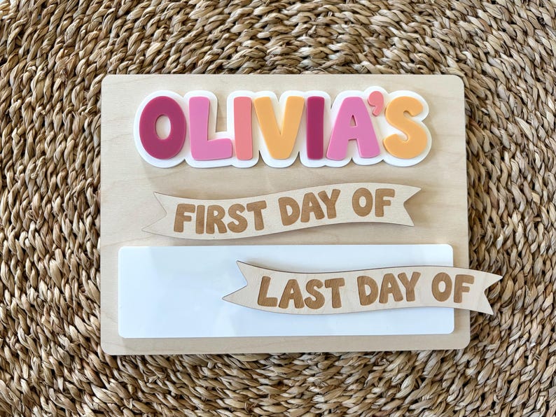 K&ouml;nnte beinhalten: Ein rechteckiges Holzschild mit dem Namen OLIVIA'S in erhabenen, bunten Buchstaben. Unter dem Namen sind zwei h&ouml;lzerne Banner-Ausschnitte angebracht, eines mit der Aufschrift FIRST DAY OF und das andere mit LAST DAY OF, &uuml;ber einer wei&szlig;en Platte.