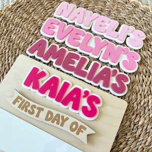 K&ouml;nnte beinhalten: Ein Holzschild mit gestapelten, geschichteten Acryl-Namensausschnitten in verschiedenen Rosa-T&ouml;nen von hell bis dunkel. Die Namen NAYELI'S, EVELYN'S, AMELIA'S und KAIA'S befinden sich &uuml;ber einem h&ouml;lzernen Banner mit der Aufschrift FIRST DAY OF.