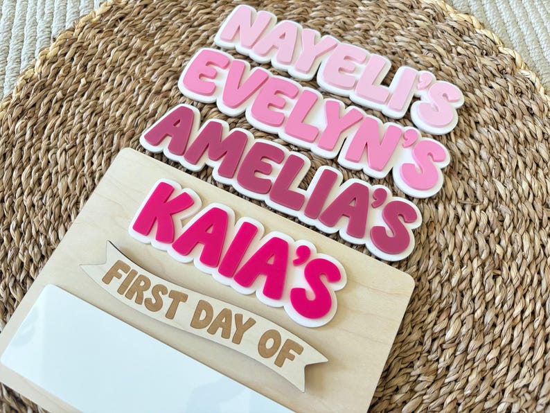 K&ouml;nnte beinhalten: Ein Holzschild mit gestapelten, geschichteten Acryl-Namensausschnitten in verschiedenen Rosa-T&ouml;nen von hell bis dunkel. Die Namen NAYELI'S, EVELYN'S, AMELIA'S und KAIA'S befinden sich &uuml;ber einem h&ouml;lzernen Banner mit der Aufschrift FIRST DAY OF.