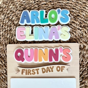K&ouml;nnte beinhalten: Ein Holzschild mit den Schriftz&uuml;gen ARLO'S, ELINA'S und QUINN'S in bunten, geschichteten Buchstaben im Bubble-Stil. Unter diesen Namen befindet sich ein Holzbanner mit dem Text FIRST DAY OF &uuml;ber einer leeren, wei&szlig;en rechteckigen Fl&auml;che.