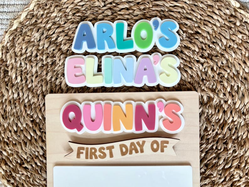 K&ouml;nnte beinhalten: Ein Holzschild mit den Schriftz&uuml;gen ARLO'S, ELINA'S und QUINN'S in bunten, geschichteten Buchstaben im Bubble-Stil. Unter diesen Namen befindet sich ein Holzbanner mit dem Text FIRST DAY OF &uuml;ber einer leeren, wei&szlig;en rechteckigen Fl&auml;che.