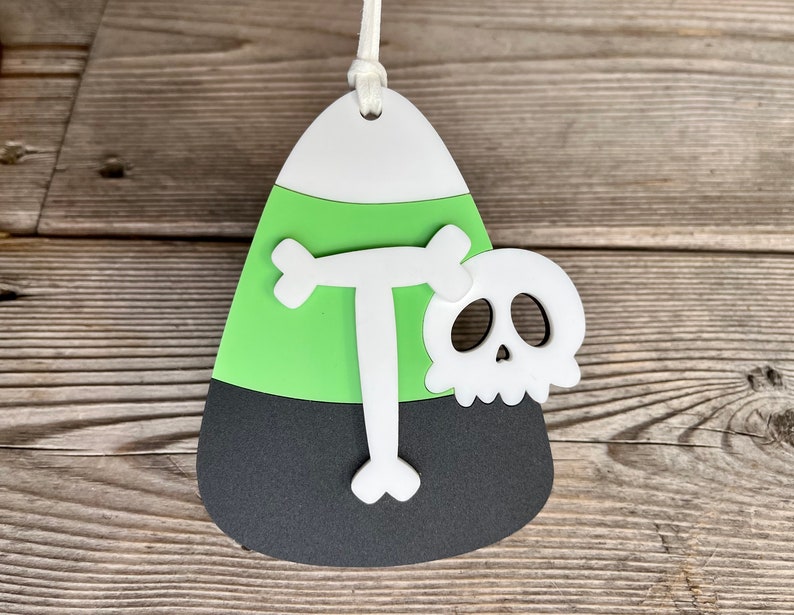 May include: A green, black, and white candy corn ornament with a white letter "T" and a white skull with black eye sockets.