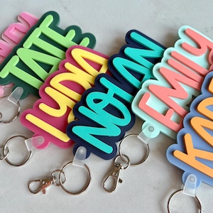 May include: A collection of six custom acrylic keychains featuring the names TATE, LYNA, NOYAN, EMILY, and KADEN in various bright colors including pink, green, yellow, dark blue, teal, and orange, each attached to a silver metal lobster clasp.