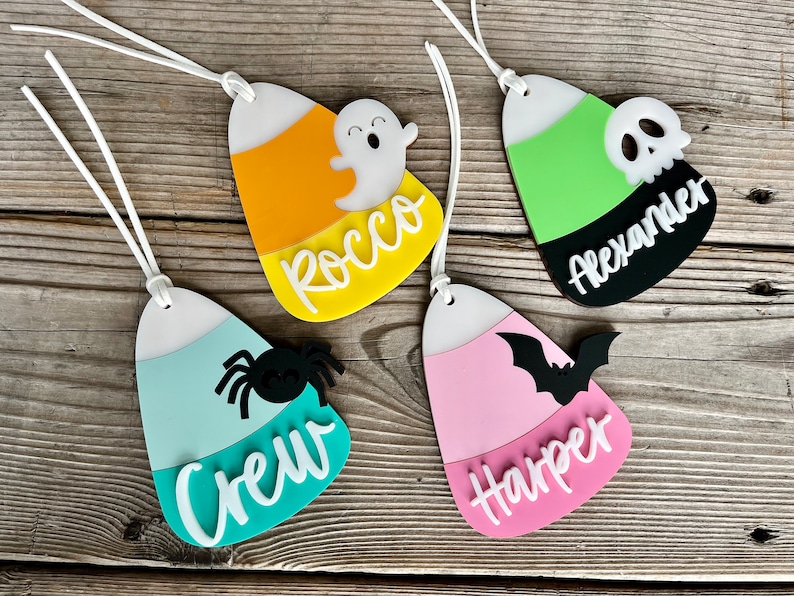 May include: Four Halloween-themed candy corn ornaments with white string hangers. The ornaments are made of acrylic and feature different Halloween characters and names: Rocco with a ghost, Crew with a spider, Harper with a bat, and Alexander with a skull.