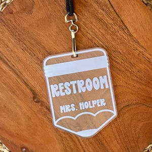 May include: A clear acrylic rectangular badge with rounded bottom corners, featuring white printed text that reads RESTROOM and MRS. HOLPER. The badge is attached to a black lanyard by a silver-toned metal swivel hook and a split ring.