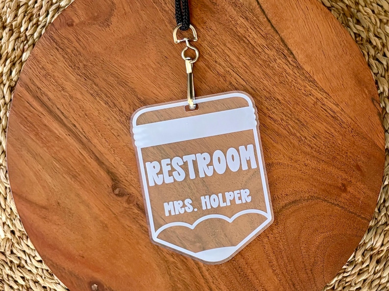 May include: A clear acrylic rectangular badge with rounded bottom corners, featuring white printed text that reads RESTROOM and MRS. HOLPER. The badge is attached to a black lanyard by a silver-toned metal swivel hook and a split ring.
