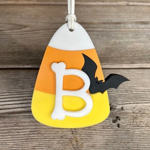 May include: A white, orange, and yellow candy corn ornament with a white letter "B" and a black bat.