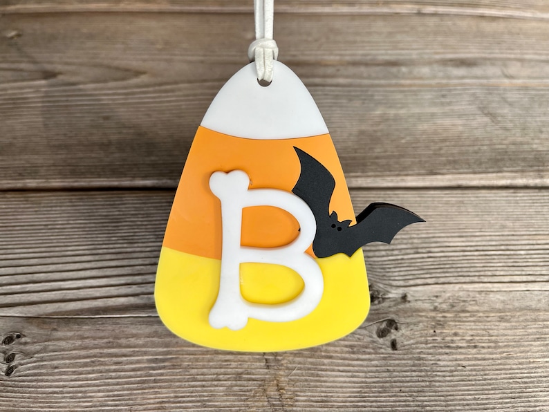 May include: A white, orange, and yellow candy corn ornament with a white letter "B" and a black bat.