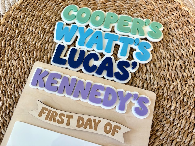 K&ouml;nnte beinhalten: Ein Holzschild mit gestapelten, farbigen 3D-Buchstaben, die die Namen Cooper's, Wyatt's, Lucas und Kennedy's bilden, &uuml;ber einem Holzbanner mit der eingravierten Aufschrift FIRST DAY OF, alles auf einer hellen Holzplatte angebracht.