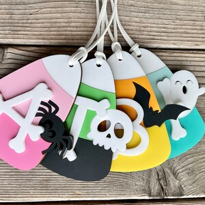 May include: Five Halloween-themed candy corn ornaments. The ornaments are made of felt and feature a variety of designs, including a spider, a skull, a bat, a ghost, and the letter "A".