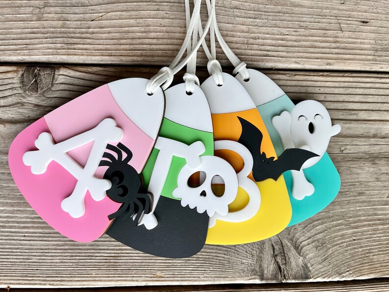 May include: Five Halloween-themed candy corn ornaments. The ornaments are made of felt and feature a variety of designs, including a spider, a skull, a bat, a ghost, and the letter "A".