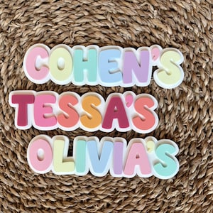 K&ouml;nnte beinhalten: Drei personalisierte Namensschilder mit den Namen Cohen's, Tessa's und Olivia's in einer fetten, abgerundeten, mehrfarbigen Schriftart. Jeder Buchstabe ist erhaben und zeigt eine Vielzahl von Pastell- und leuchtenden Farben wie Rosa, Gelb, Blau und Lila.