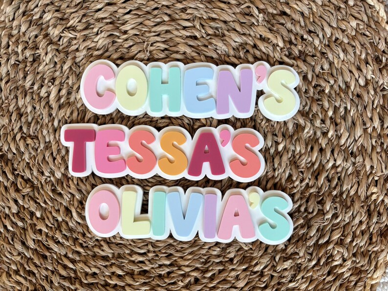 K&ouml;nnte beinhalten: Drei personalisierte Namensschilder mit den Namen Cohen's, Tessa's und Olivia's in einer fetten, abgerundeten, mehrfarbigen Schriftart. Jeder Buchstabe ist erhaben und zeigt eine Vielzahl von Pastell- und leuchtenden Farben wie Rosa, Gelb, Blau und Lila.