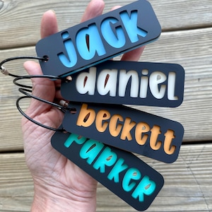 May include: Four rectangular black luggage tags with cut-out lettering revealing colorful inserts. The tags feature the names Jack in light blue, Daniel in white, Beckett in orange, and Parker in teal. Each tag includes a black wire loop fastener.