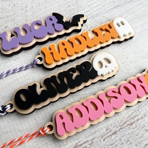 May include: Four personalized Halloween-themed wooden tags with names "LUCK", "HADLEY", "OLIVER", and "ADDISON" in colorful, wavy lettering. Each tag features a different Halloween icon: a bat, a ghost, a skull, and a pumpkin.
