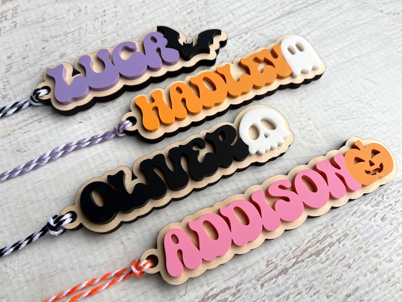 May include: Four personalized Halloween-themed wooden tags with names "LUCK", "HADLEY", "OLIVER", and "ADDISON" in colorful, wavy lettering. Each tag features a different Halloween icon: a bat, a ghost, a skull, and a pumpkin.