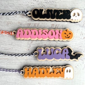 May include: Four Halloween-themed wooden name tags with string attached. The names are Oliver, Addison, Luca, and Hadley. Each tag has a different Halloween icon: skull, pumpkin, bat, and ghost.