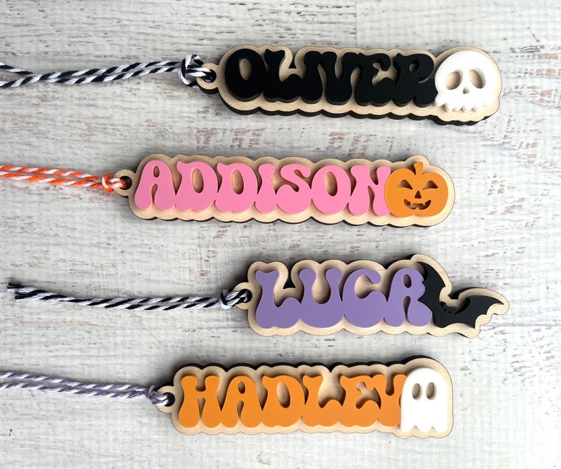 May include: Four Halloween-themed wooden name tags with string attached. The names are Oliver, Addison, Luca, and Hadley. Each tag has a different Halloween icon: skull, pumpkin, bat, and ghost.