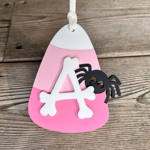 May include: Pink and white candy corn shaped ornament with a white letter A and a black spider on the side.