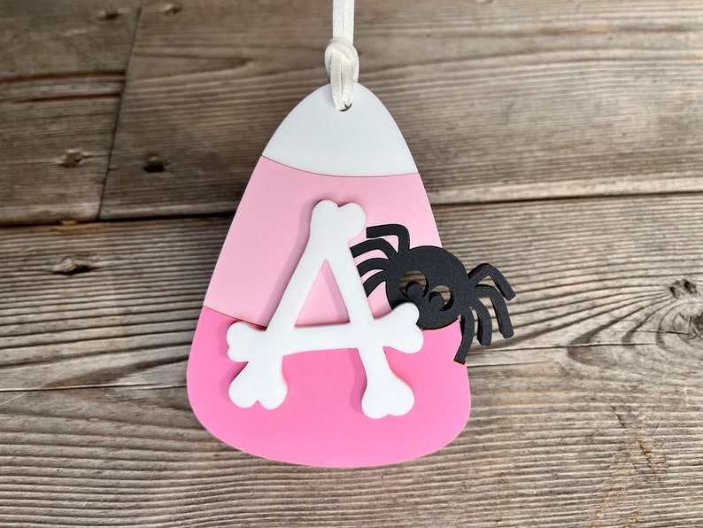 May include: Pink and white candy corn shaped ornament with a white letter A and a black spider on the side.