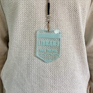 May include: A light blue acrylic badge holder shaped like a pencil, hanging from a black lanyard. The badge features the text LIBRARY and MRS. HOLPER in a bold font, with a pencil tip design at the bottom, worn over a textured white sweater.