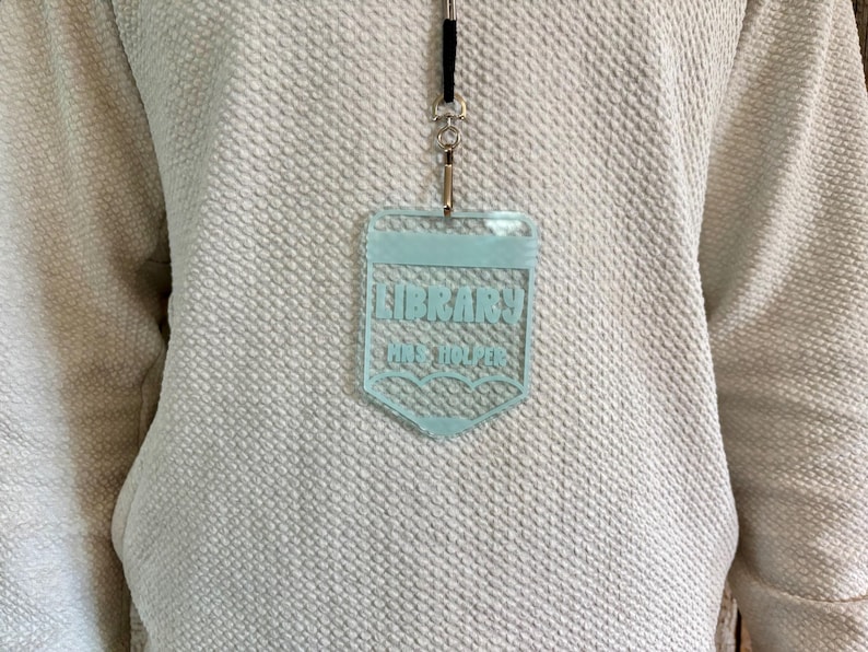 May include: A light blue acrylic badge holder shaped like a pencil, hanging from a black lanyard. The badge features the text LIBRARY and MRS. HOLPER in a bold font, with a pencil tip design at the bottom, worn over a textured white sweater.