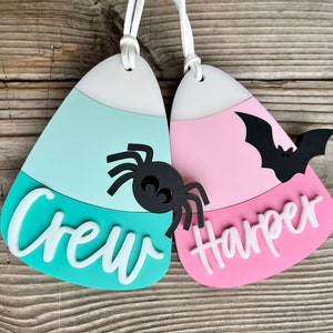 May include: Two candy corn shaped Halloween ornaments with white string hangers. The teal ornament has a black spider and the text "Crew" in white. The pink ornament has a black bat and the text "Harper" in white.