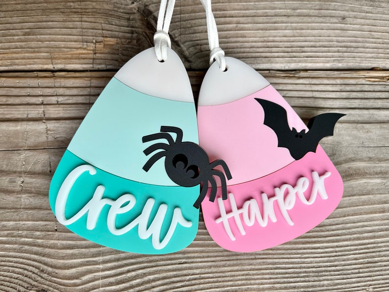 May include: Two candy corn shaped Halloween ornaments with white string hangers. The teal ornament has a black spider and the text "Crew" in white. The pink ornament has a black bat and the text "Harper" in white.