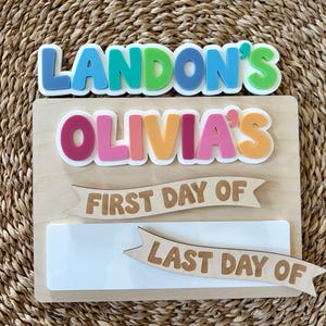 K&ouml;nnte beinhalten: Ein rechteckiges Holzschild mit bunten 3D-Buchstaben, die LANDON'S und OLIVIA'S lesen. Unter den Namen befinden sich zwei Holzbanner mit dem Text FIRST DAY OF und LAST DAY OF &uuml;ber einer leeren wei&szlig;en rechteckigen Fl&auml;che zum Beschriften.