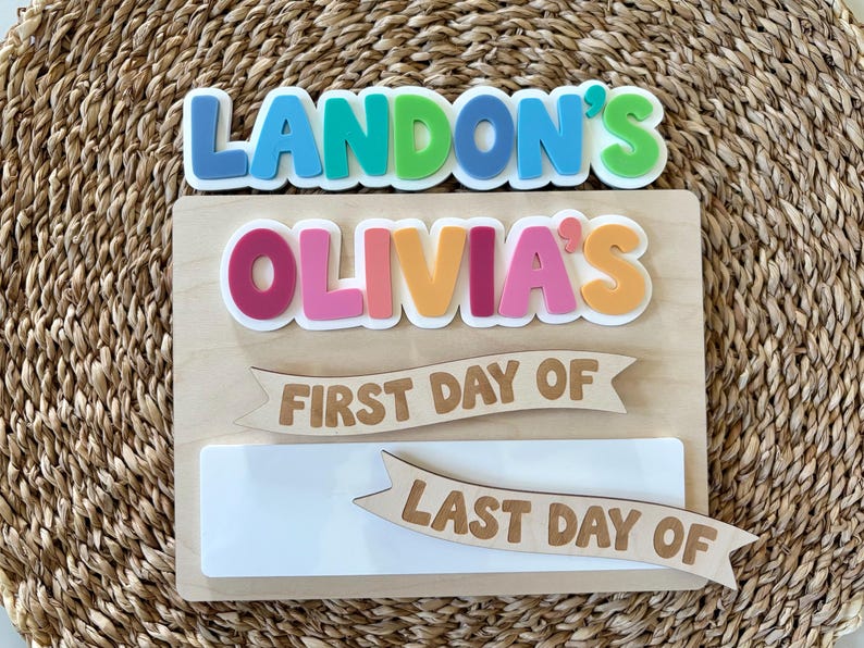 K&ouml;nnte beinhalten: Ein rechteckiges Holzschild mit bunten 3D-Buchstaben, die LANDON'S und OLIVIA'S lesen. Unter den Namen befinden sich zwei Holzbanner mit dem Text FIRST DAY OF und LAST DAY OF &uuml;ber einer leeren wei&szlig;en rechteckigen Fl&auml;che zum Beschriften.