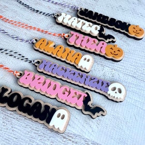 May include: Seven Halloween-themed wooden ornaments with names written in colorful, stylized lettering. Each ornament features a different Halloween icon, including a ghost, a bat, a skull, and a pumpkin.