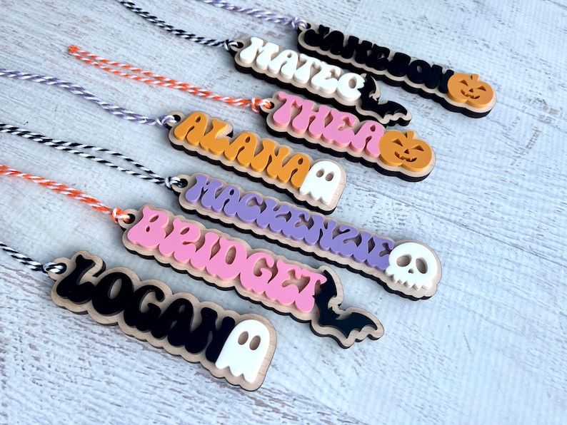 May include: Seven Halloween-themed wooden ornaments with names written in colorful, stylized lettering. Each ornament features a different Halloween icon, including a ghost, a bat, a skull, and a pumpkin.