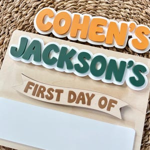 K&ouml;nnte beinhalten: Ein Holzschild mit den Namen COHEN'S und JACKSON'S in erhabener orangefarbener und dunkelgr&uuml;ner Schrift. Unter den Namen zeigt ein Banner den Text FIRST DAY OF in Braun, gefolgt von einem leeren wei&szlig;en rechteckigen Feld zum Beschriften.