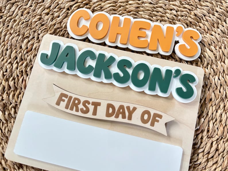 K&ouml;nnte beinhalten: Ein Holzschild mit den Namen COHEN'S und JACKSON'S in erhabener orangefarbener und dunkelgr&uuml;ner Schrift. Unter den Namen zeigt ein Banner den Text FIRST DAY OF in Braun, gefolgt von einem leeren wei&szlig;en rechteckigen Feld zum Beschriften.