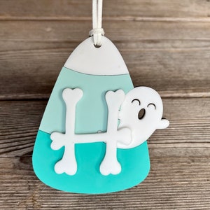 May include: A teal and white candy corn shaped ornament with a white ghost and a white bone shaped letter H.