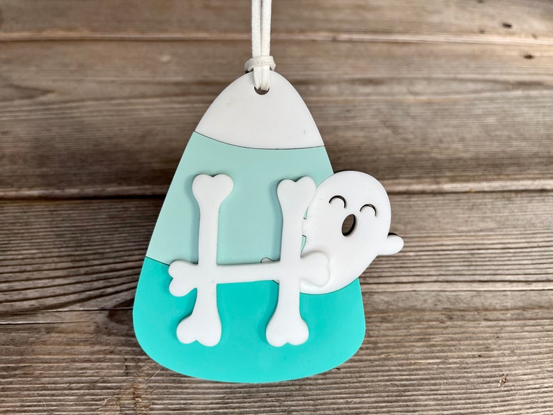 May include: A teal and white candy corn shaped ornament with a white ghost and a white bone shaped letter H.
