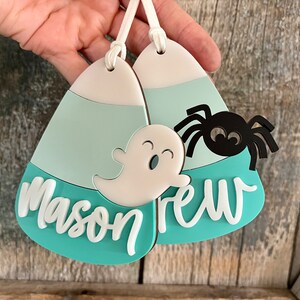 May include: Two teal blue and white candy corn shaped ornaments with a ghost and spider design. The ornaments are personalized with the names "Mason" and "Drew".