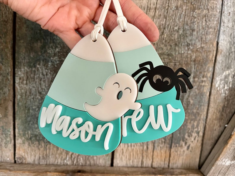 May include: Two teal blue and white candy corn shaped ornaments with a ghost and spider design. The ornaments are personalized with the names "Mason" and "Drew".