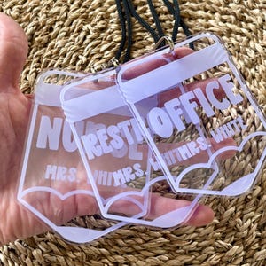 May include: A clear acrylic badge holder shaped like a pencil with a black lanyard. The holder features white text that reads REST OFFICE and MRS. WHITMRS. WHITE. The design includes a white pencil tip outline at the bottom of the clear plastic.