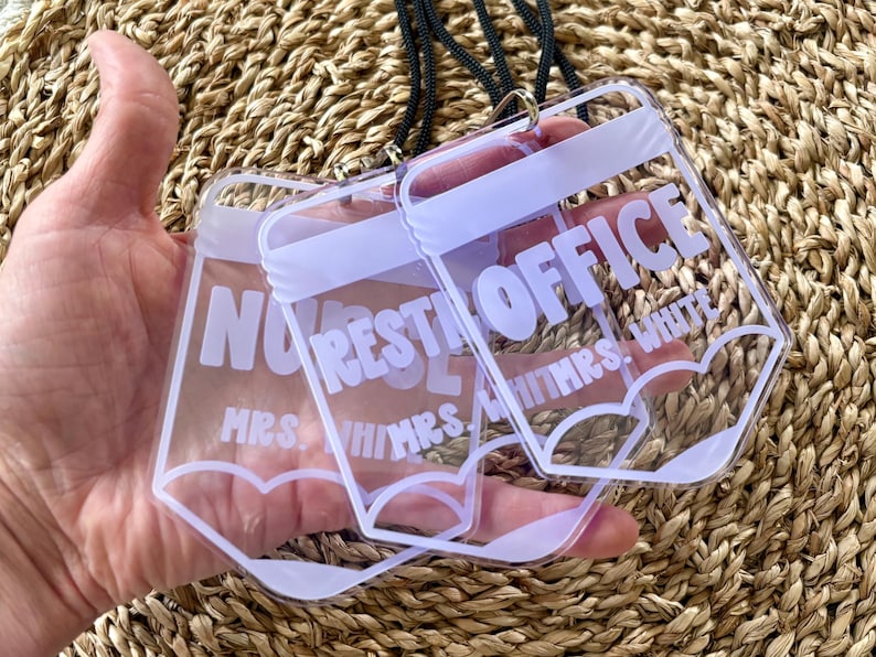 May include: A clear acrylic badge holder shaped like a pencil with a black lanyard. The holder features white text that reads REST OFFICE and MRS. WHITMRS. WHITE. The design includes a white pencil tip outline at the bottom of the clear plastic.