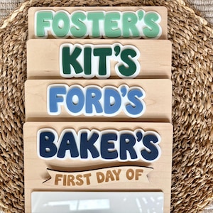 K&ouml;nnte beinhalten: Ein Stapel aus vier rechteckigen Holzschildern mit heller Maserung. Jedes Schild hat erhabene, farbige Schriftz&uuml;ge mit den Namen FOSTER'S, KIT'S, FORD'S und BAKER'S. Das unterste Schild enth&auml;lt ein Holzbanner mit dem Text FIRST DAY OF.