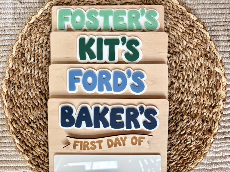 K&ouml;nnte beinhalten: Ein Stapel aus vier rechteckigen Holzschildern mit heller Maserung. Jedes Schild hat erhabene, farbige Schriftz&uuml;ge mit den Namen FOSTER'S, KIT'S, FORD'S und BAKER'S. Das unterste Schild enth&auml;lt ein Holzbanner mit dem Text FIRST DAY OF.