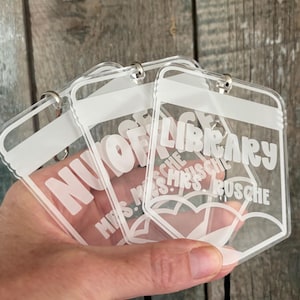 May include: Three clear acrylic pencil-shaped tags with white printed text. The tags feature the word LIBRARY at the top and the name MRS. RUSCHE below, with a pencil tip graphic at the bottom and a metal ring attached to the top of each tag.