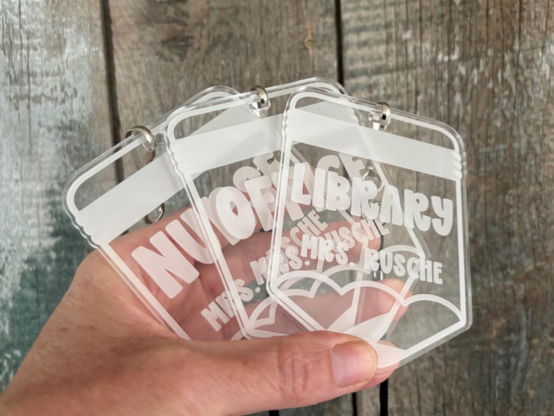 May include: Three clear acrylic pencil-shaped tags with white printed text. The tags feature the word LIBRARY at the top and the name MRS. RUSCHE below, with a pencil tip graphic at the bottom and a metal ring attached to the top of each tag.