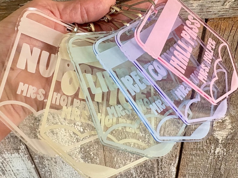 May include: A set of four rectangular clear acrylic keychains with rounded corners, each featuring a pastel-colored border and printed text. The text on the keychains includes the words HALL PASS and MRS. HOLPER, along with heart-shaped graphics.