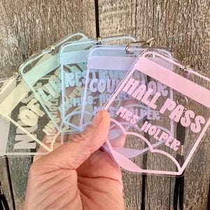 May include: A set of clear acrylic rectangular hall passes with rounded corners, each featuring pastel-colored text and a matching lanyard. The passes display various labels including Hall Pass, Library, and Restroom, along with the name Mrs. Holper.